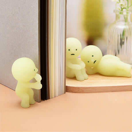 Three small, yellow, glow-in-the-dark plastic figurines from Smiski Series 1—featuring humanoid shapes where one holds a book, another sits, and the third reclines on a wooden surface—evoke curiosity much like treasures revealed from their blind box.