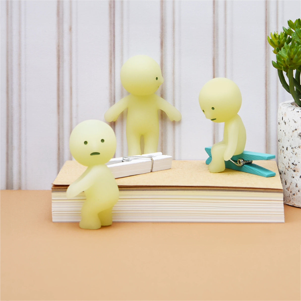 A trio of small, pale green Smiski Series 1 figurines are thoughtfully positioned on and around a stack of books. One of the Glow-in-the-Dark figures from the Smiski Series 1 - Blind Box is perched atop a large paper clip. A small plant can be seen in the background, enhancing the scene's mysterious charm.