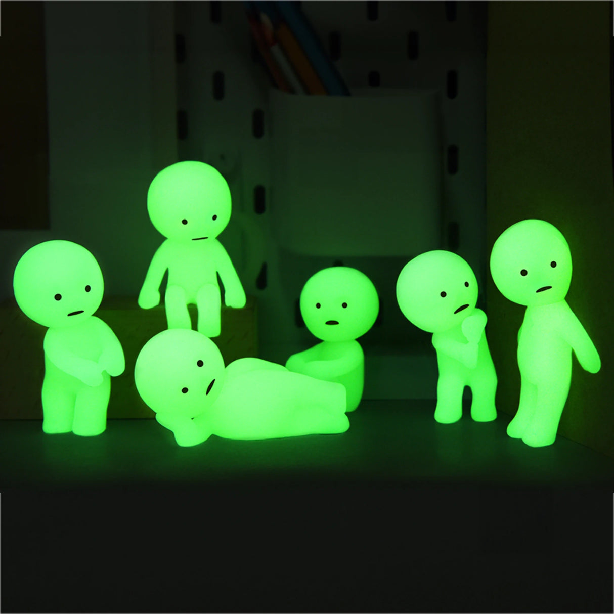 Against a dark backdrop, six small, glowing green figures resembling humans from Smiski's Series 1 take on various poses. These charming figures, packaged in Smiski's Blind Boxes, enchant with their captivating glow-in-the-dark feature and add a whimsical touch.