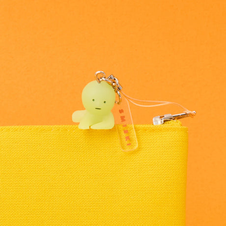 A Smiski Strap Accessory Keychain - Series 2 by Smiski, resembling a small green collectible figure, is attached to the zipper of a yellow pouch, shown against an orange background.