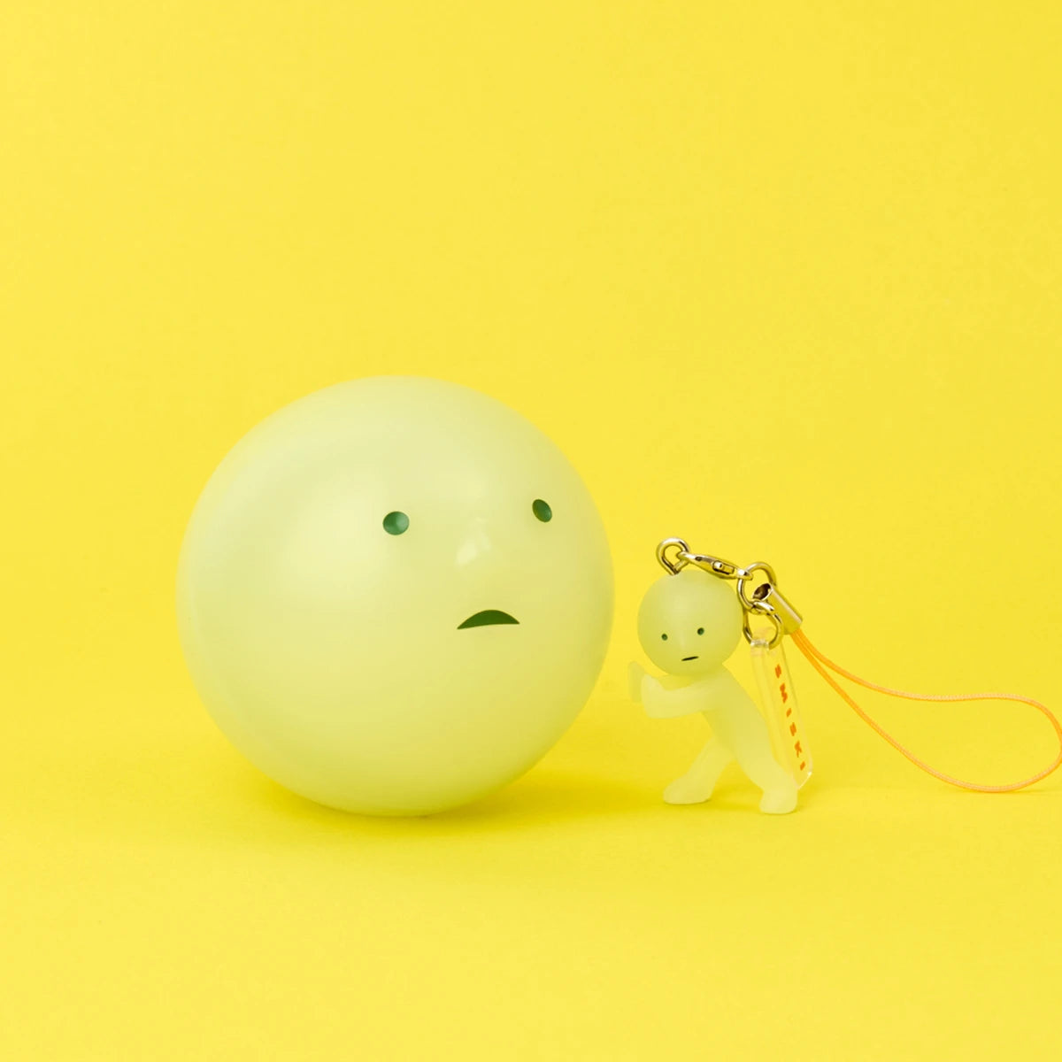 A Smiski Strap Accessory Keychain - Series 2 by Smiski stands beside a larger round object; both display simple, expressionless faces on a yellow background.