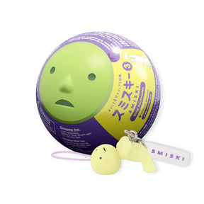 Small pale yellow character keychain lying down with white tag next to round purple and yellow capsule toy container
