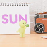 A Smiski Sunday Series - Blind Box figurine glows on a table holding a green object. In the background, a white notepad with "SUN" in purple and a vintage camera create an intriguing scene.