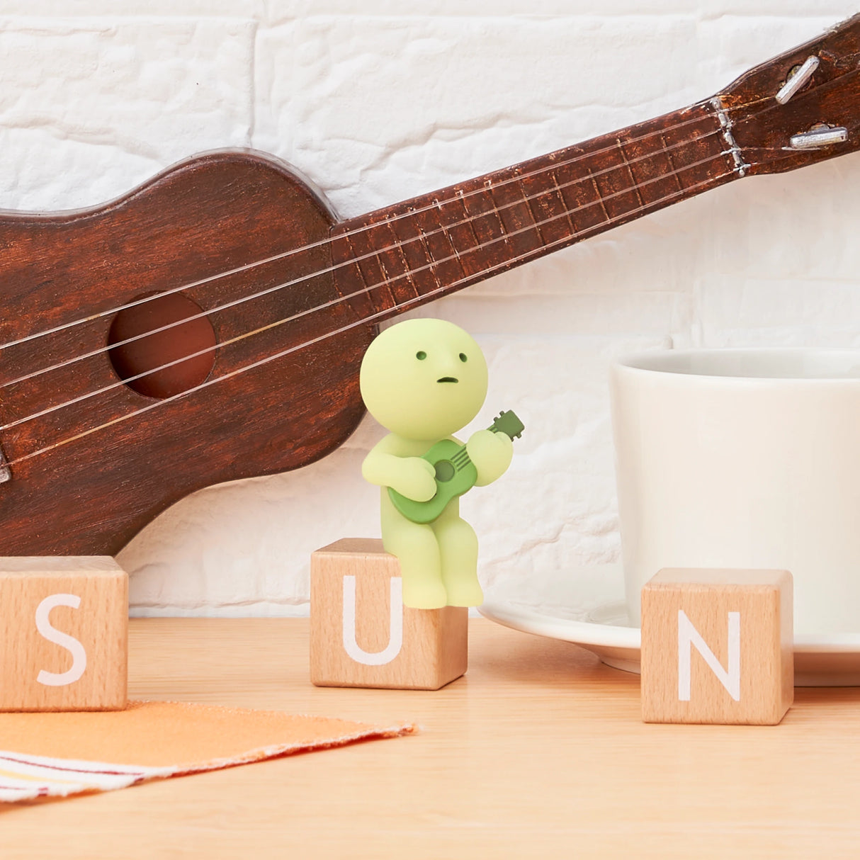 From the Smiski Sunday Series by Smiski, a small green figure glows in the dark while holding a ukulele and sitting on wooden blocks spelling "SUN" beside a coffee cup, with a larger ukulele resting against the white brick wall.