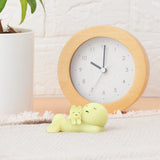 A small, glow-in-the-dark figure from the Smiski Sunday Series and a cat relax on a beige mat. A round wooden clock and a white plant pot complete the cozy scene, embodying the charm of Smiski.