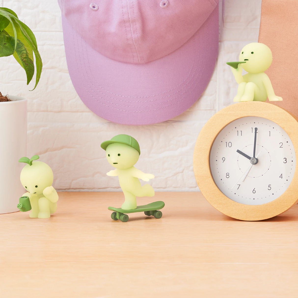 Three small glow-in-the-dark figurines from the Smiski Sunday Series - Blind Box are on a desk: one with a leaf, one on a skateboard, and one with a straw hat. They sit beside a clock, potted plant, and pink cap hanging on the wall.