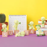 Smiski Sunday Series figures feature activities like eating, guitar playing, skateboarding, and relaxing by calendar, cactus, and books on "SUNDAY" blocks against a yellow background. Some glow in the dark! Each Blind Box offers a surprise from Smiski.