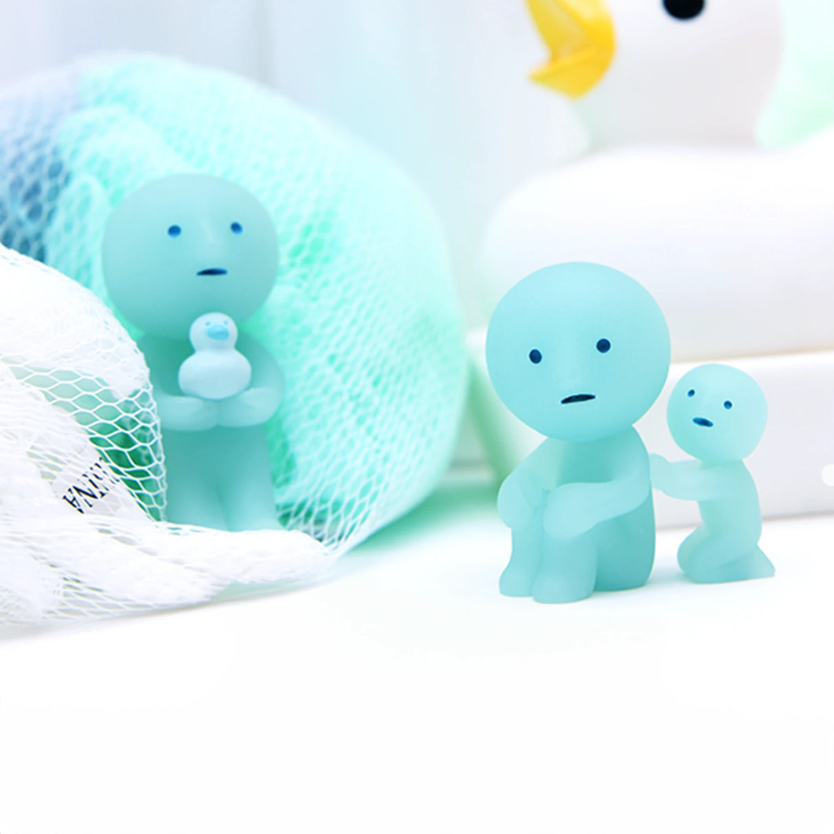 A small blue figure from the Smiski Bath Series - Blind Box, featuring a child inside a mesh bag, stands next to another figure holding a smaller one. In the background, a large rubber duck looms while glow-in-the-dark collectible figures from Smiski add an enchanting touch.