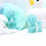 A small blue figure from the Smiski Bath Series - Blind Box, featuring a child inside a mesh bag, stands next to another figure holding a smaller one. In the background, a large rubber duck looms while glow-in-the-dark collectible figures from Smiski add an enchanting touch.