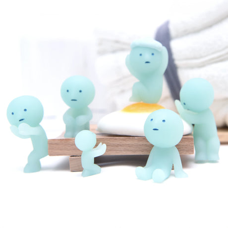 Six Smiski Bath Series - Blind Box figurines, featuring a small, blue, ghost-like design and various expressions, gather around a fried egg model on a wooden platform. They glow in the dark amidst stacked towels, bringing a whimsical touch to any bathroom setting.