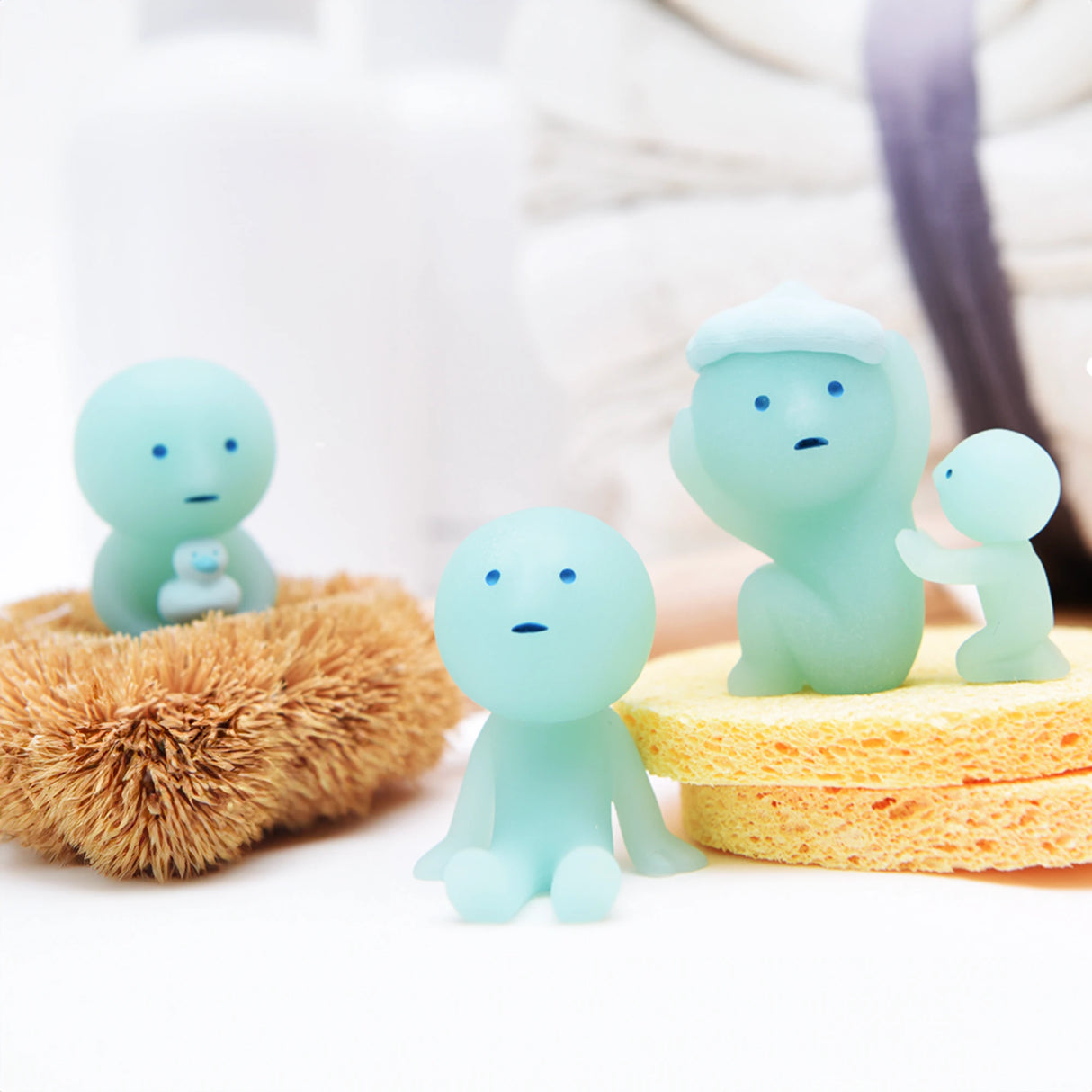 Three small figurines from the Smiski Bath Series - Blind Box, all in blue, sit and stand on bath sponges. One holds a glow-in-the-dark green baby, another perches in a brush, with a stack of towels behind them. These adorable collectibles from Smiski bring charm to any bathroom setting.