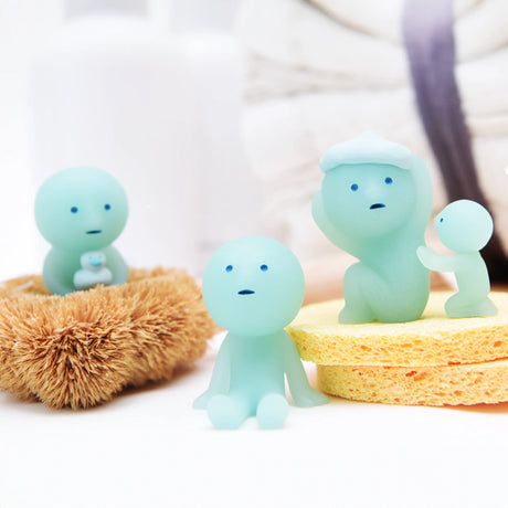 Three small figurines from the Smiski Bath Series - Blind Box, all in blue, sit and stand on bath sponges. One holds a glow-in-the-dark green baby, another perches in a brush, with a stack of towels behind them. These adorable collectibles from Smiski bring charm to any bathroom setting.