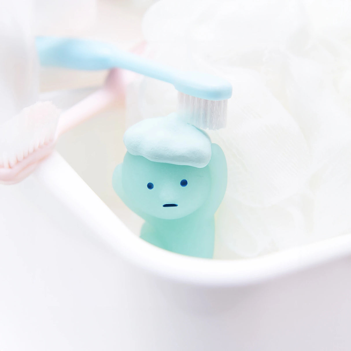 A charming Smiski figurine from the Bath Series Blind Box, featuring a small blue figure with a face, stands in a white container alongside two toothbrushes and white fabric. This delightful collectible from the Smiski brand glows in the dark, bringing a unique touch to its serene setting.