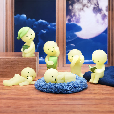 Six glow-in-the-dark, yellow figurines in various poses with books create a whimsical scene. Known as the Smiski Bed Series - Blind Box by Smiski, these bedtime buddies sit by a window showcasing a nighttime sky with a full moon, adding charm to any evening routine.