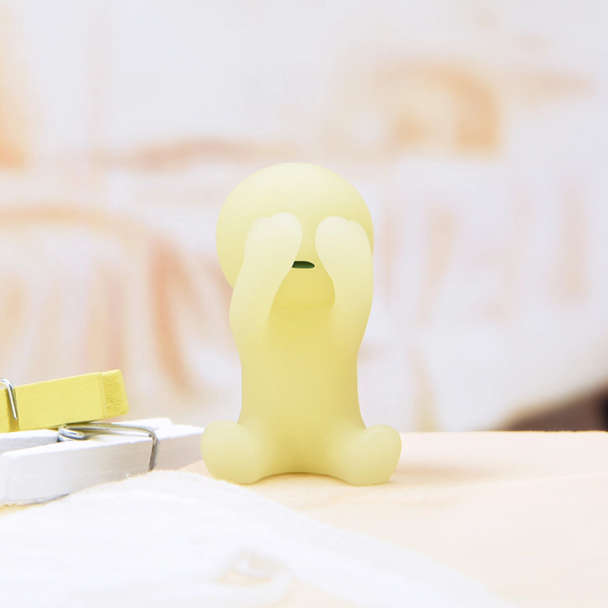 A small, smooth, yellow figure from the Smiski Bed Series - Blind Box by Smiski is perched on a surface with its hands covering its eyes. These delightful figures make perfect bedtime companions, glowing in the dark to keep night fears away.