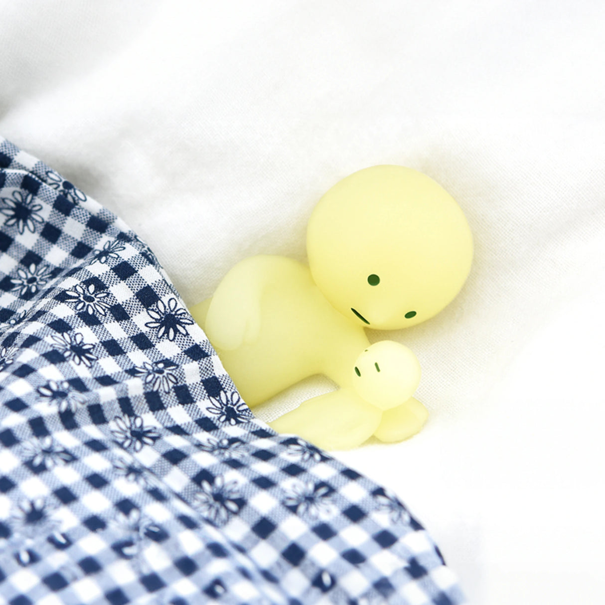 Two round, yellow figures with simple facial features lie on a white surface partially covered by a blue and white checkered cloth with floral patterns, resembling glowing bedtime buddies from the Smiski Bed Series - Blind Box by Smiski.