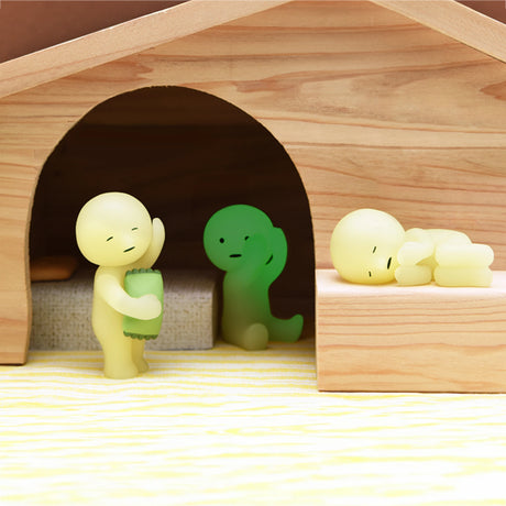 Inside a wooden structure, three small, abstract figurines from the Smiski Bed Series - Blind Box by Smiski are arranged. One figurine stands while holding an object, another sits, and the third lies down with a glow-in-the-dark feature that adds a restful touch. These charming bedtime companions enhance any space with their whimsical poses and gentle luminescence.