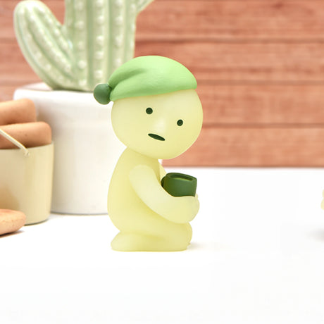 A petite glow-in-the-dark green figure from the Smiski Bed Series - Blind Box by Smiski clutches a cup while sitting on a white surface, with a cactus plant and jar in the backdrop. This charming little companion adds coziness and fun to your bedtime routine.