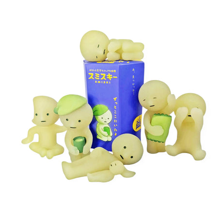 Six small, pale green Smiski figurines, each in different poses and with glow-in-the-dark features, encircle a blue and purple box adorned with Japanese text. As part of the Smiski Bed Series - Blind Box collection by Smiski, these enchanting bedtime companions bring a whimsical charm to any space.