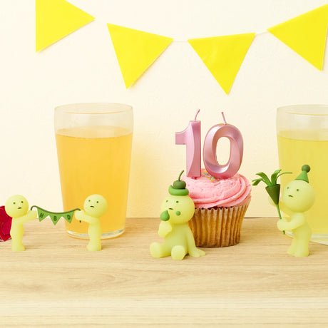 Two glasses of juice, a pink cupcake with a "10" candle, and Smiski Birthday Series - Blind Box figurines by Smiski holding decorations sit on a table with a yellow pennant banner in the background.