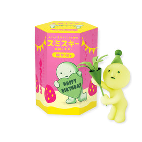 A small yellow Smiski Birthday Series figurine with a party hat holds a plant beside the colorful Smiski Birthday Series - Blind Box featuring Japanese text and cake illustrations.