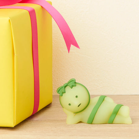 The Smiski Birthday Series - Blind Box by Smiski features a small green Glow-in-the-Dark figurine wrapped in green ribbons with a bow, lying next to a yellow gift box with a pink ribbon on a wooden surface.
