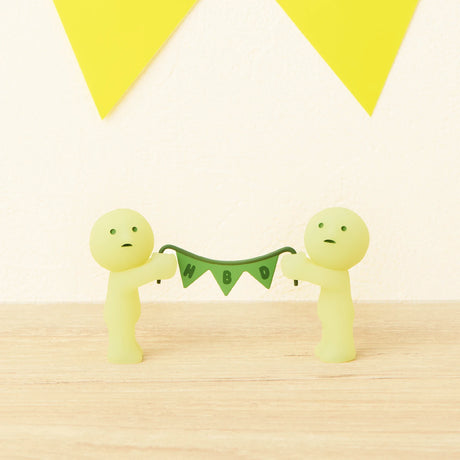 Two small, pale green Smiski figurines from the Smiski Birthday Series - Blind Box hold a green "HBD" pennant banner, with yellow triangular flags in the background.