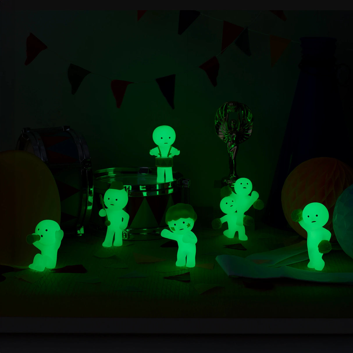 The Smiski Cheer Series – Blind Box glow-in-the-dark figurines by Smiski are artfully arranged on a table, surrounded by vibrant party decorations and a small drum. These charming figures add a mysterious touch to any celebration.