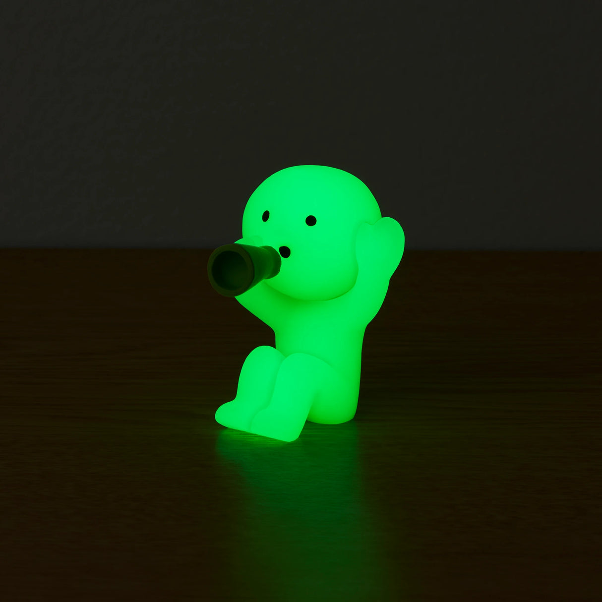 A small figure from the Smiski Cheer Series - Blind Box by Smiski sits on a dark surface, holding a tiny bottle to its mouth. This glow-in-the-dark character captivates with its mysterious luminescence.