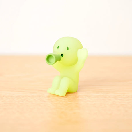 A tiny figure from the Smiski Cheer Series - Blind Box holds a nozzle to its mouth on a wooden surface, glowing softly in green.