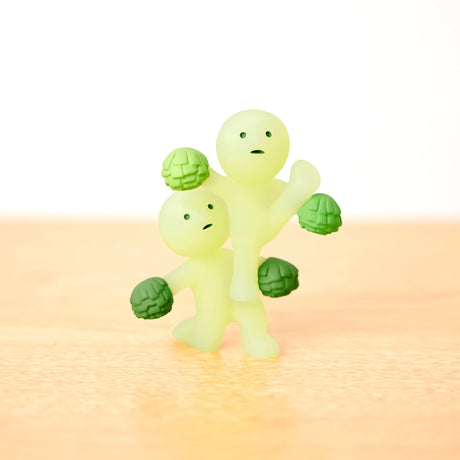 Two pale green, round-headed figures with small eyes and mouths stand on a wooden surface, each featuring four green pom-pom-like hands. These glow-in-the-dark figures from the Smiski Cheer Series - Blind Box add an element of surprise for those who enjoy a blind box experience.