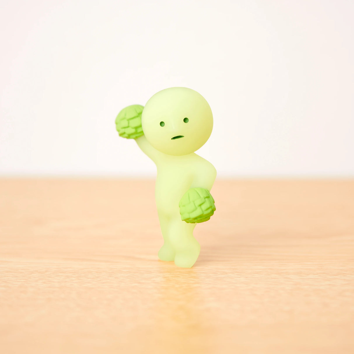 A small, round-headed figurine from the Smiski Cheer Series - Blind Box collection holds two green items while standing on a wooden surface against a plain background. Part of the charming glow-in-the-dark figures from Smiski, it brings an unexpected twist to your display.