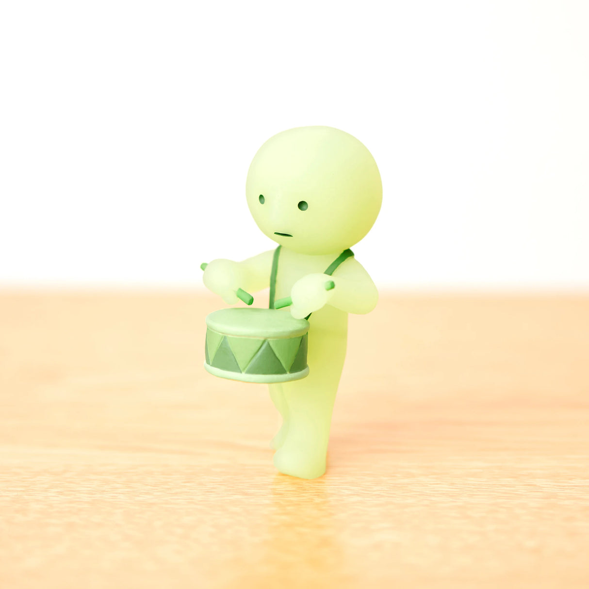 A small green Smiski figurine from the Cheer Series Blind Box is playing a miniature drum, standing on a light beige surface.