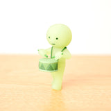 A small green Smiski figurine from the Cheer Series Blind Box is playing a miniature drum, standing on a light beige surface.