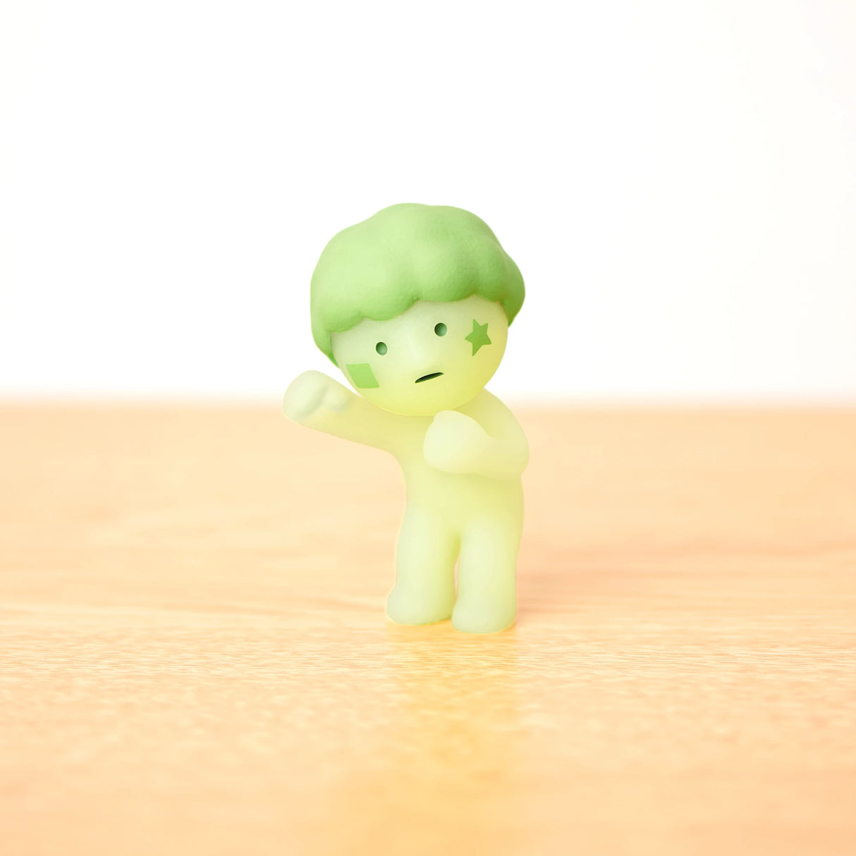 The Smiski Cheer Series - Blind Box by Smiski features a glow-in-the-dark figure with a green star on its cheek and hair, striking a fighting pose on a light wooden surface. Experience the excitement—each blind box holds a surprise.