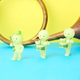 Three pastel green figures from the Smiski Cheer Series - Blind Box by Smiski march in a line, drums in hand, against a vibrant blue and yellow backdrop. A partial view of a French horn peeks into the scene as these glow-in-the-dark figures exude an enchanting charm.