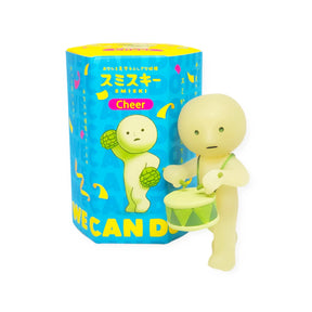 A toy figure with a drum and pom-poms is positioned beside a blue hexagonal box labeled "Cheer" from the Smiski Cheer Series, displaying a vibrant confetti pattern.