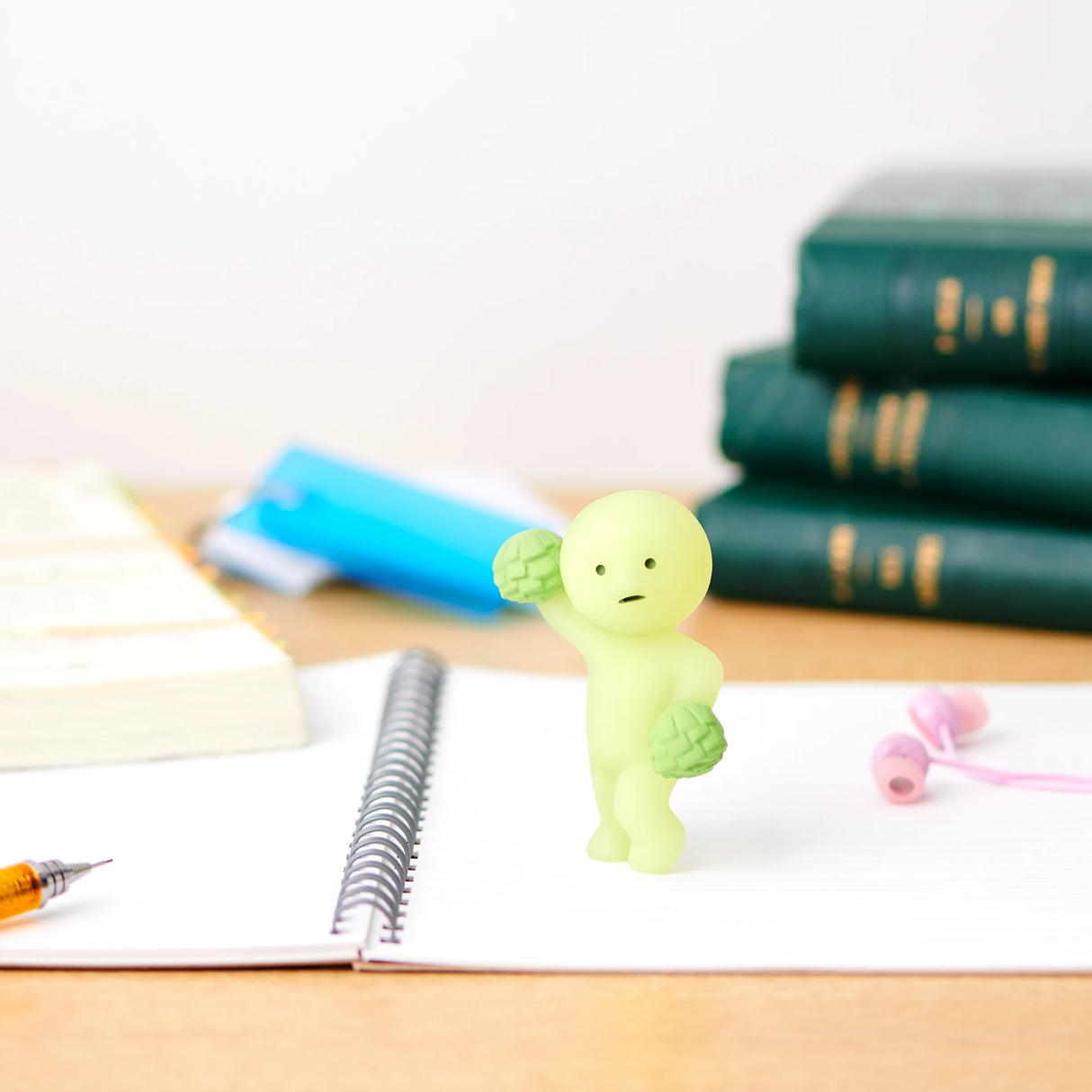 A small green figure from the Smiski Cheer Series - Blind Box by Smiski stands on an open notebook, surrounded by stacked books and earphones on the desk. These charming glow-in-the-dark figures create a whimsical atmosphere, sparking curiosity just like opening a blind box.