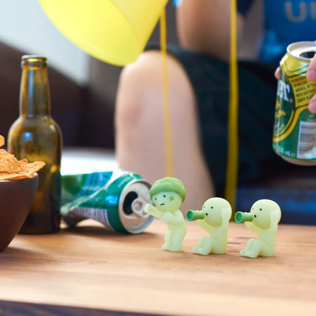 Three small figures from the Smiski Cheer Series - Blind Box are blowing horns on a coffee table surrounded by snack bowls, a beer bottle, and crumpled soda cans. Someone nearby is holding a can. These glow-in-the-dark figures by Smiski add a whimsical touch to the casual scene.