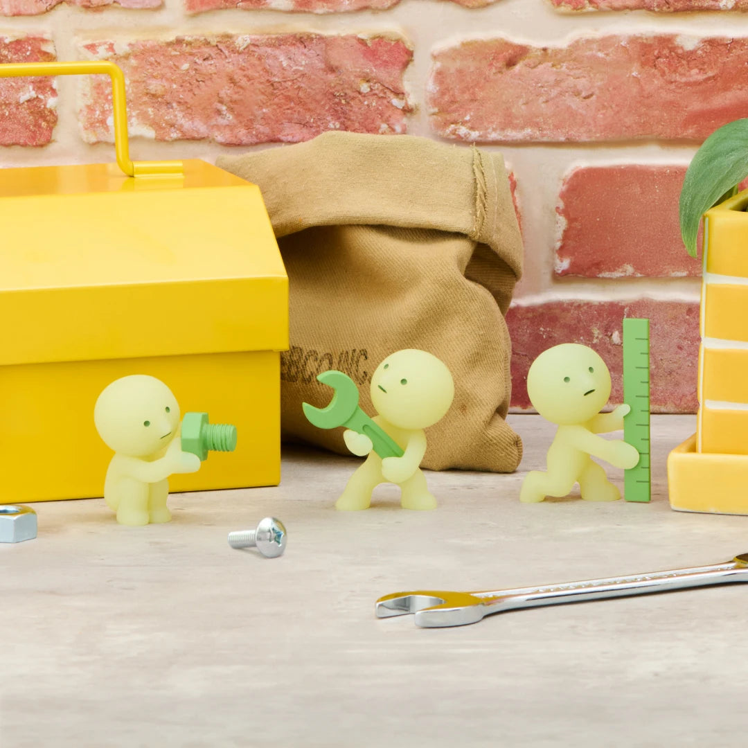 Smiski construction figures in front of yellow boxes, light green minis holding small tools