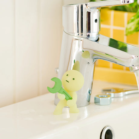 Yellow-green construction figure with a green wrench-like tool in front of a chrome faucet