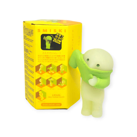 A small figurine wearing a green shirt and glowing in the dark is positioned next to a yellow box labeled "Smiski Dressing Series - Blind Box," which showcases illustrations of similar characters from the series.