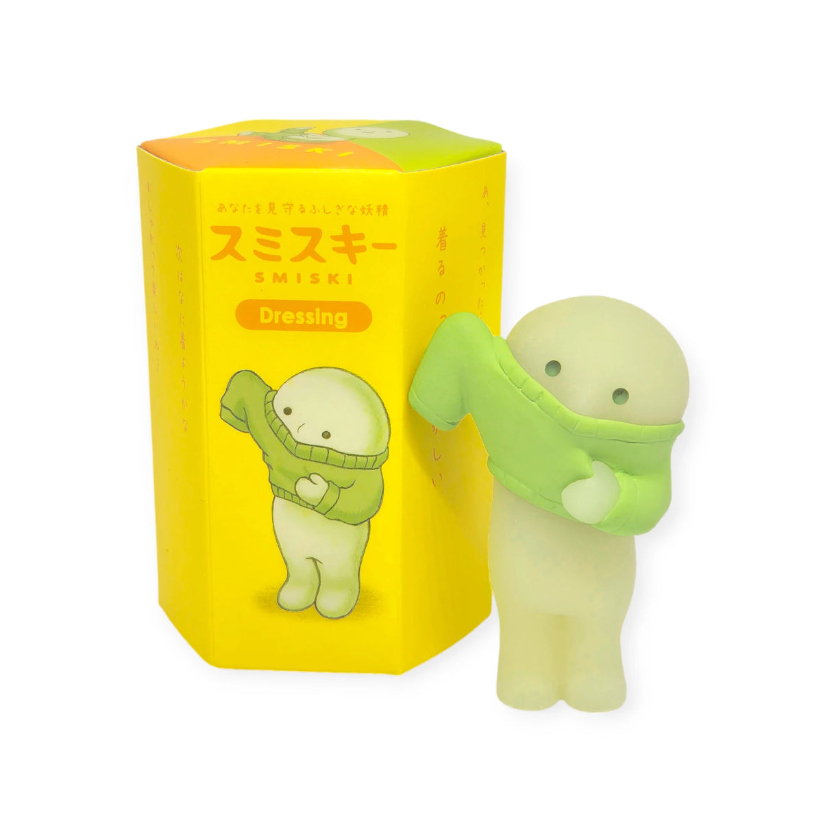 A small, glow-in-the-dark figure is holding a green piece of clothing. Next to it is a yellow box featuring Japanese text and the words "Smiski Dressing Series - Blind Box" from the Smiski brand in English.