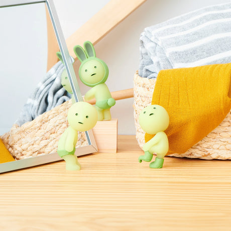 Three glow-in-the-dark Smiski Dressing Series figures, each with a unique expression, are displayed on a wooden surface. One figure gazes into a mirror, another rests against a fabric basket, and the third perches on the edge of a yellow cloth. These enchanting characters from Smiski add charm to any space.