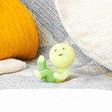 A small, green, glow-in-the-dark clay figure from the Smiski Dressing Series - Blind Box by Smiski features a round head and is seated on a textured surface, surrounded by white and yellow cushions, adding an enchanting touch to any space.