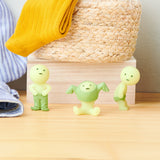 Three figures from the Smiski Dressing Series - Blind Box, by the brand Smiski, stand on a wooden table next to a woven basket and a yellow cloth. These small, green humanoid figures glow in the dark, adding an intriguing charm to the scene.