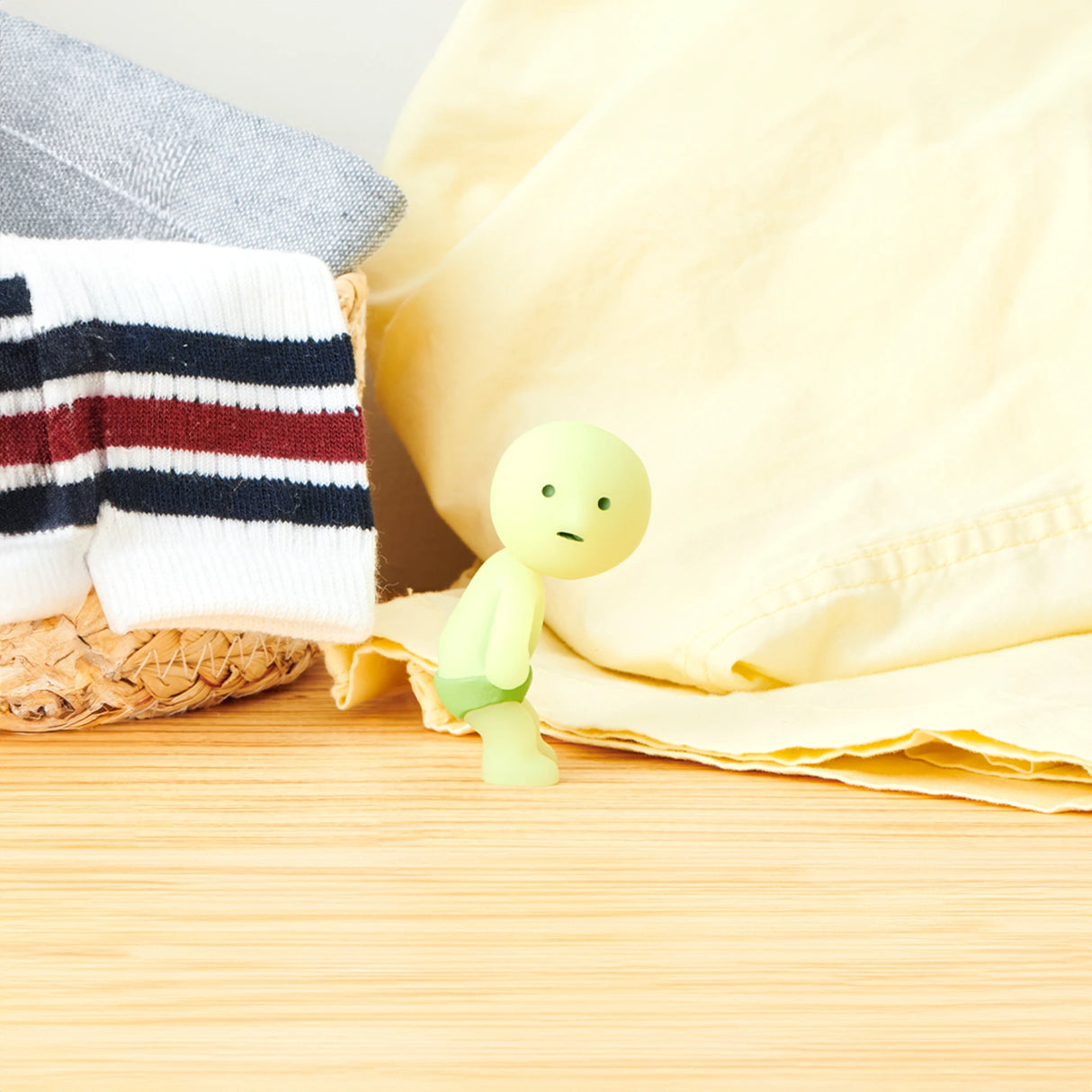 A small green figure from the Smiski Dressing Series - Blind Box by Smiski, exhibiting a concerned expression, is positioned beside a laundry basket containing socks and a yellow blanket on a wooden surface. These glow-in-the-dark figures provide an intriguing touch to any environment.