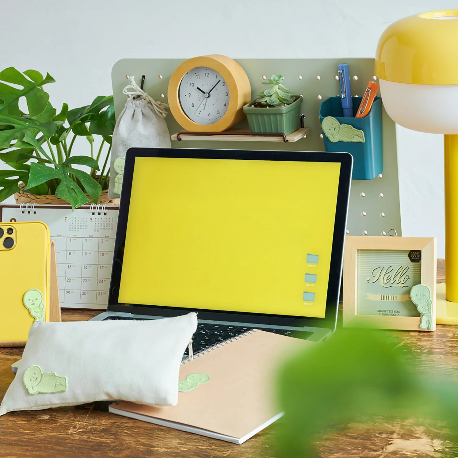 A workspace featuring a yellow laptop, green plants, clock, lamp, calendar, stationery, and the Smiski Embroidery Sticker/Patch by Smiski adds charm as an embroidered accessory on the wooden desk.