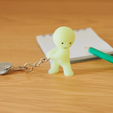 A Smiski Keychain - Carrying Key Figure from the Smiski brand is pulling a chain beside a notepad and green pencil on a wooden surface.