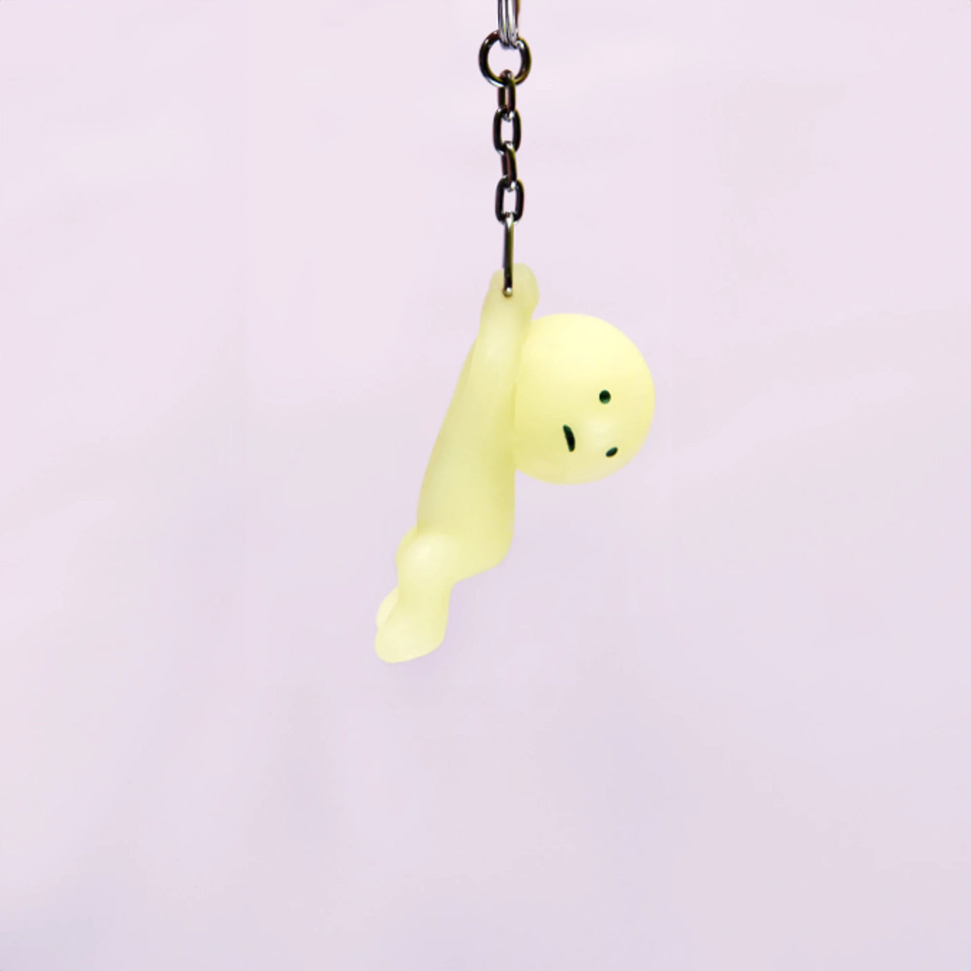 The adorable Smiski Keychain - Grabbing Key Figure, with its charming yellow hue, glows in the dark as it hangs from a chain against a simple backdrop.